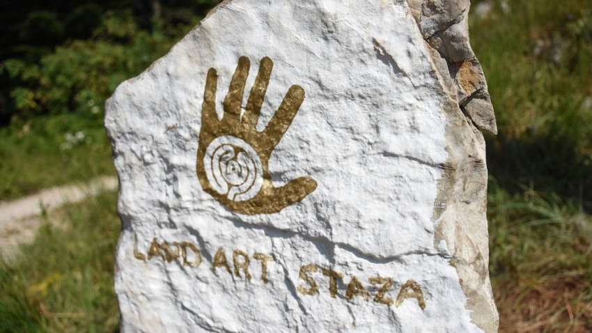 Land art trail on Učka, a stone signpost with a palm print in golden color and a labyrinth in the middle of the palm. It is the logo of the Land art trail. The palm print is on a white background, and under it is written LAND ART STAZA.